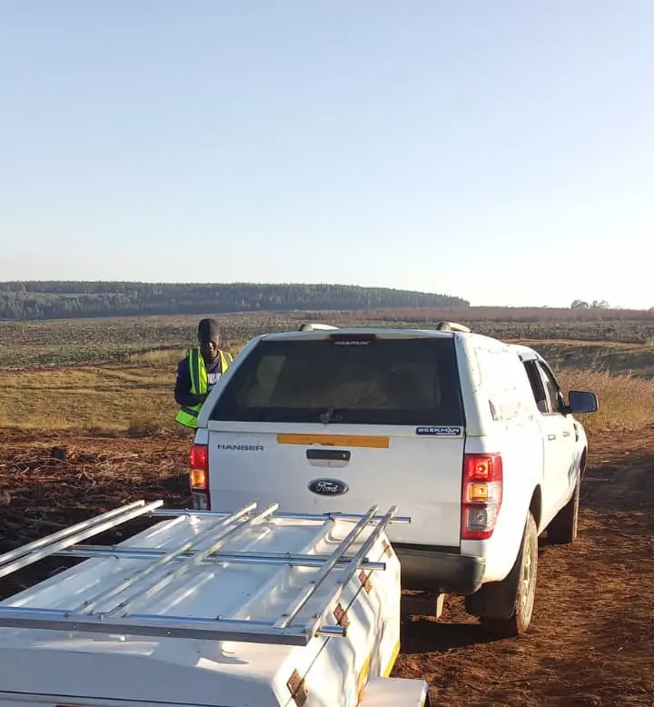 A white bakkie towing an enclosed trailer on a red-soil plantation road, with a crew member in a hi-vis vest beside it.